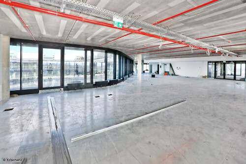 Großer, offener Büroraum im Rohbau mit Betonboden, sichtbaren Leitungen und raumhohen Fenstern.