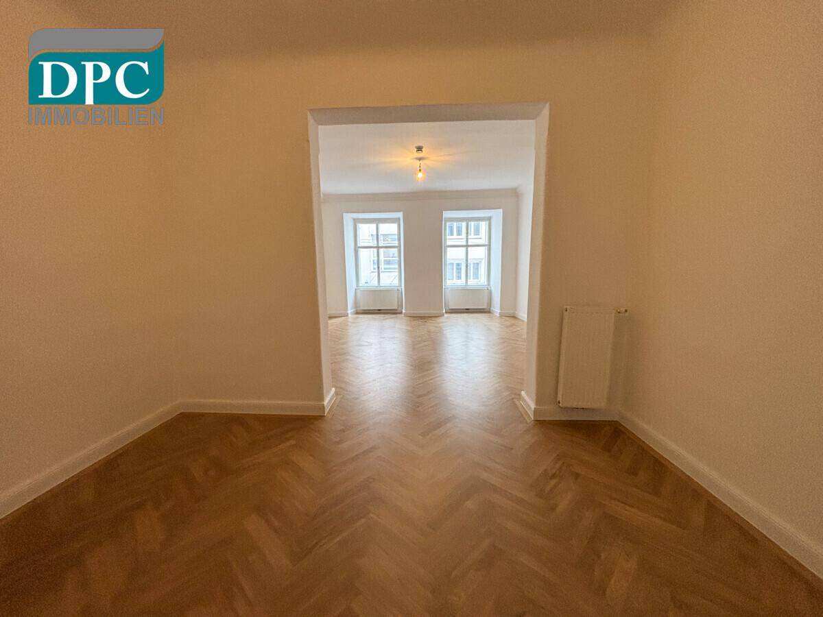 Heller Raum mit Fischgrätparkett und einem breiten Durchgang zu einem weiteren Zimmer mit zwei Fenstern.