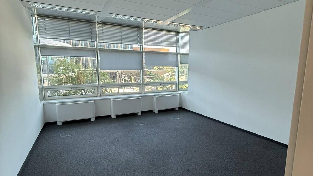 Heller Büroraum mit Teppichboden, weißen Wänden und einer Fensterfront mit Jalousien.