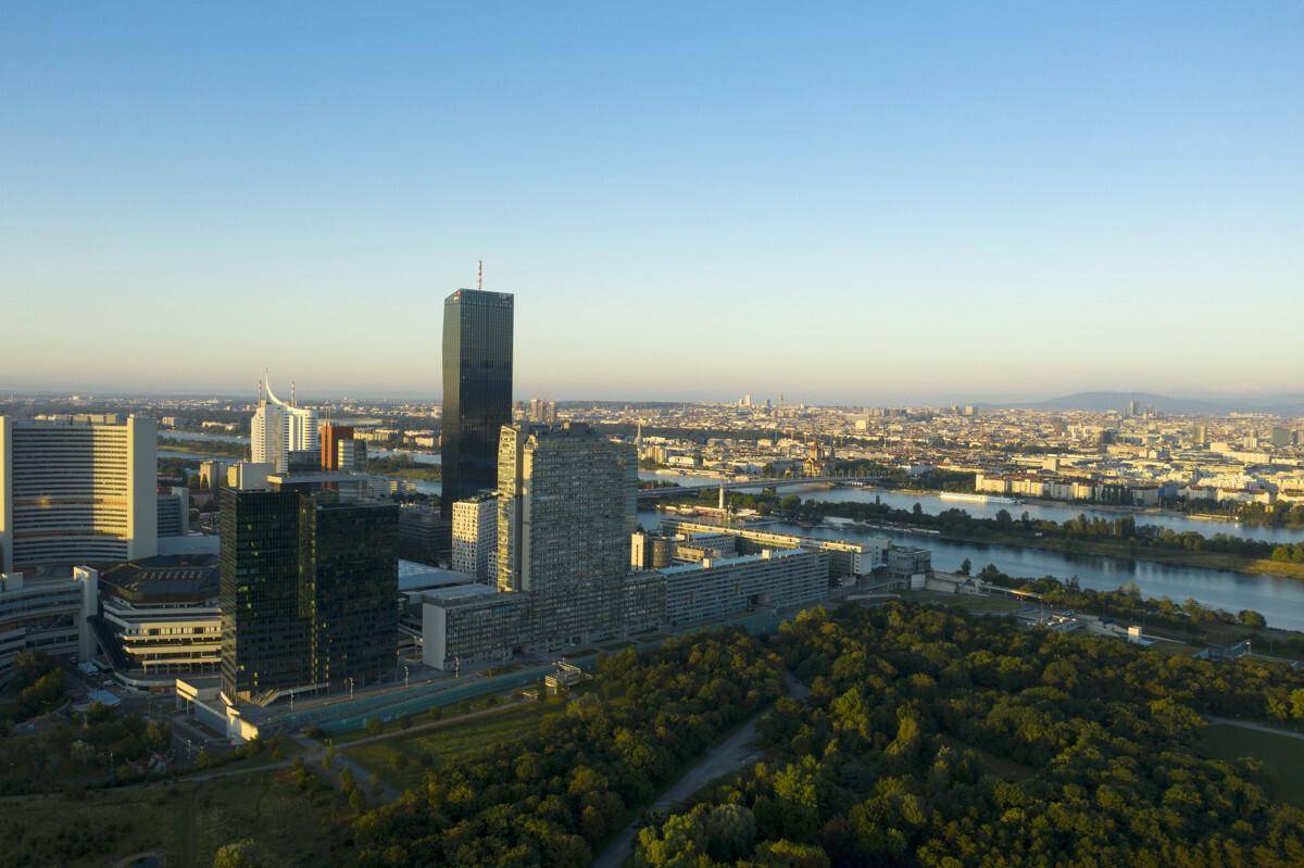 Weitläufiger Luftbildblick auf eine Stadt mit Fluss, Hochhäusern und ausgedehnten Grünflächen bei Sonnenaufgang.