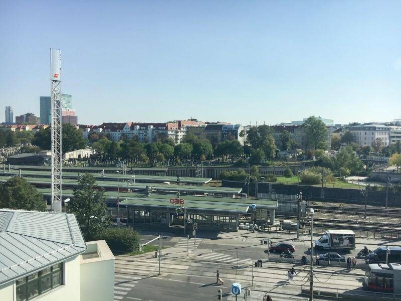 Weitläufiger Blick über eine Stadtlandschaft mit Bahngleisen, einem Bahnhof und umliegenden Gebäuden unter blauem Himmel.