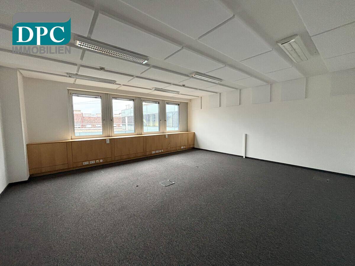 Geräumiger Büroraum mit Teppichboden, Fenstern und schallabsorbierenden Deckenplatten für angenehme Akustik.