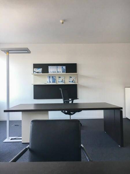 Einzelner Büroschreibtisch mit Stuhl und Wandregal in einem hellen Raum mit Teppichboden.