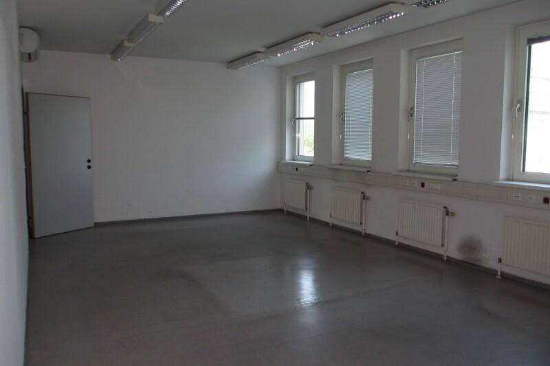 Leeres Büro mit hellen Wänden, grauem Boden und mehreren Fenstern, die viel Tageslicht hereinlassen.