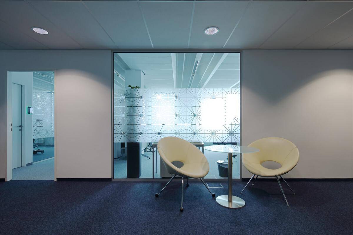Gemütlicher Wartebereich im Büro mit zwei modernen Sesseln und einem Glastisch vor einer gemusterten Glaswand.