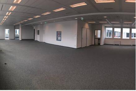 Weitläufiger, leerer Büroraum mit Teppichboden, abgehängter Decke und mehreren Türen.