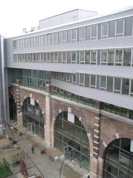 Fassade eines modernen Gebäudes mit großen Fenstern über historischen Backsteinbögen im Erdgeschoss.
