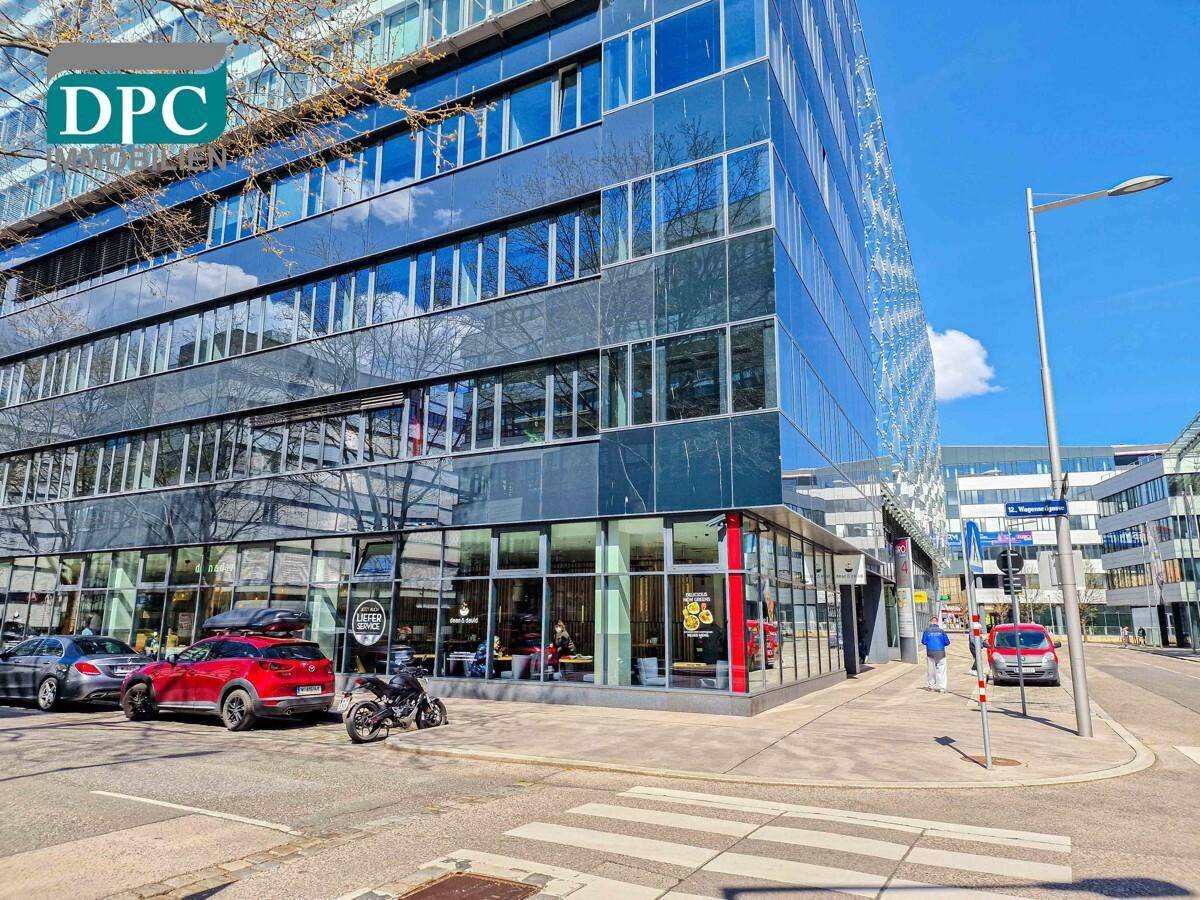 Modernes Bürogebäude mit Glasfassade an einer belebten Straße mit parkenden Autos und Fußgängern.