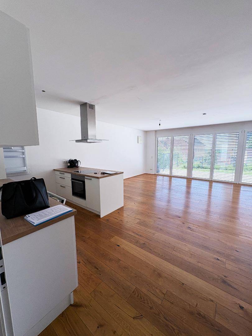 Heller Wohnbereich mit Holzboden, offener Küche und großen Fenstern mit Rollläden.