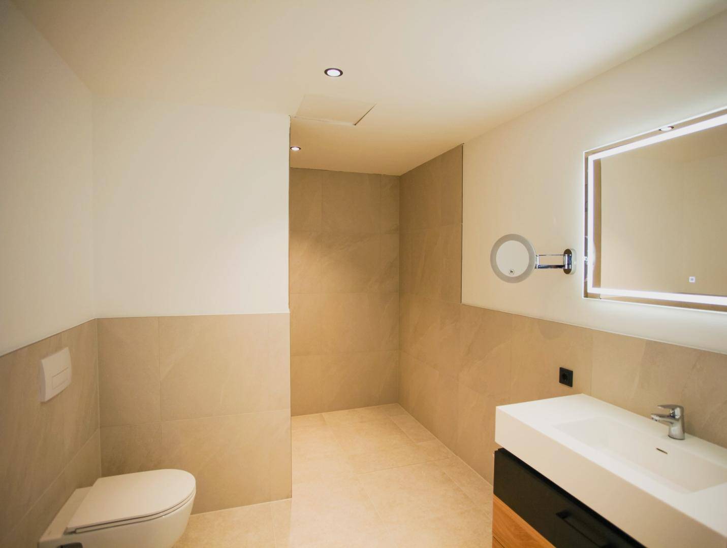 Modernes Badezimmer mit beigefarbenen Fliesen, wandhängendem WC und beleuchtetem Spiegel.