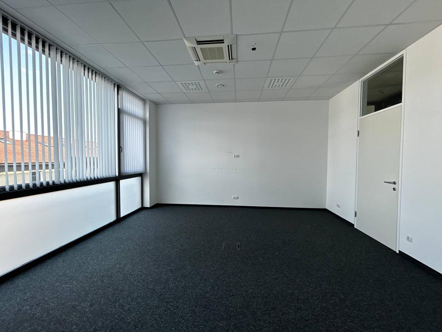 Leerer Büroraum mit großen Fenstern, vertikalen Jalousien und dunklem Teppichboden, bietet viel Tageslicht.