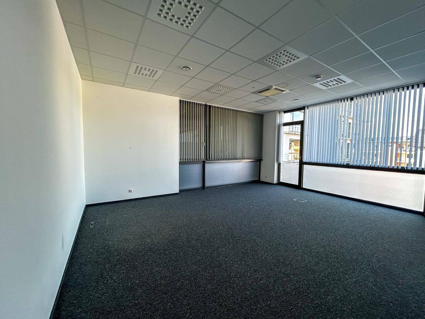Leeres Büro mit großen Fenstern, vertikalen Jalousien und dunklem Teppichboden, bereit zur individuellen Gestaltung.