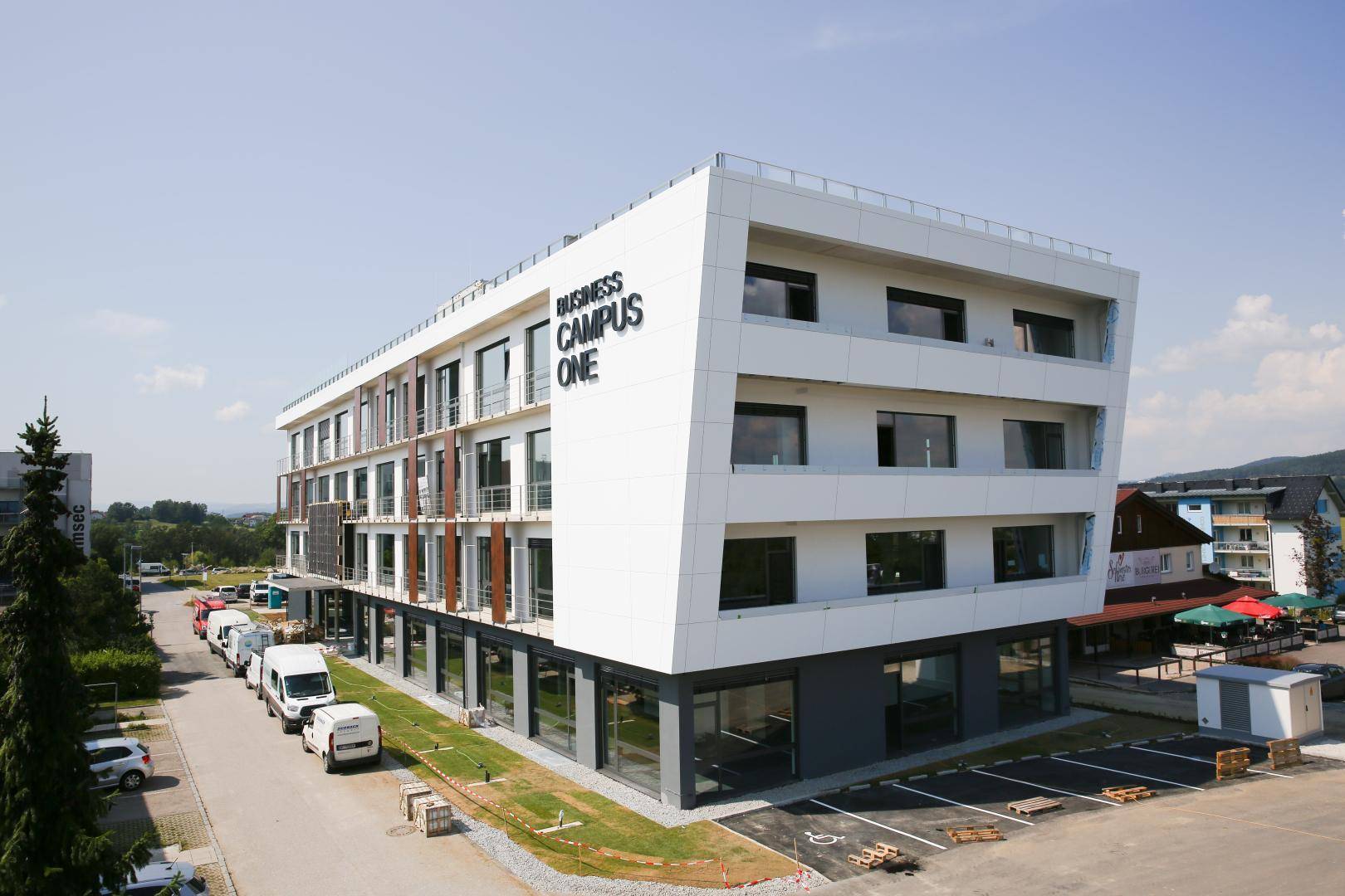 Modernes Geschäftsgebäude mit weißer Fassade, großen Fenstern und Parkplätzen unter blauem Himmel.