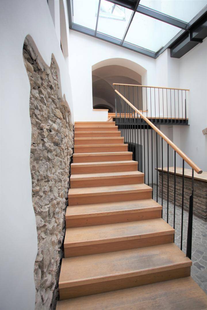 Nahaufnahme der Holztreppe und der freigelegten Steinwand, die zum hellen Glasdach führt.