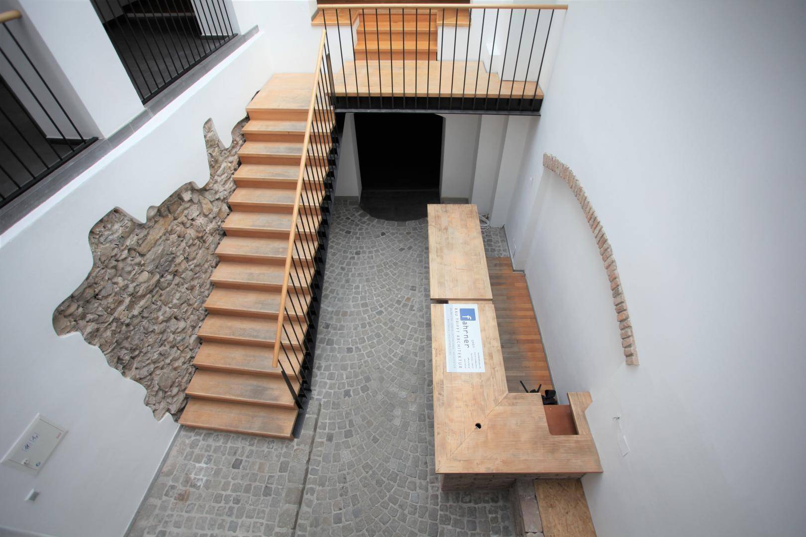 Übersicht des Eingangsbereichs mit Holztreppe, Steinwand und dem Kopfsteinpflasterboden von oben.