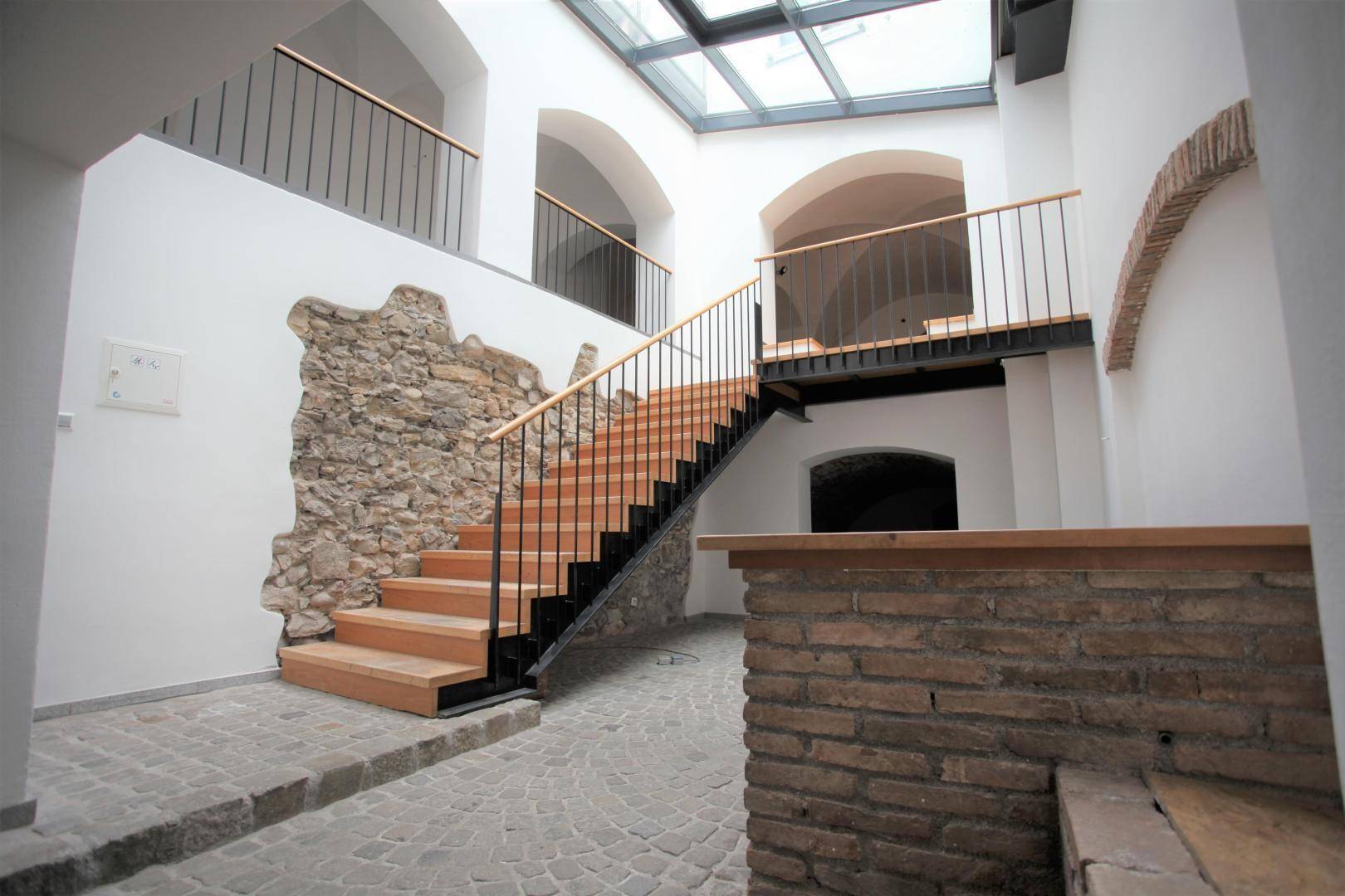 Heller Eingangsbereich mit moderner Holztreppe, freigelegter Steinwand und Glasdach für viel Tageslicht.