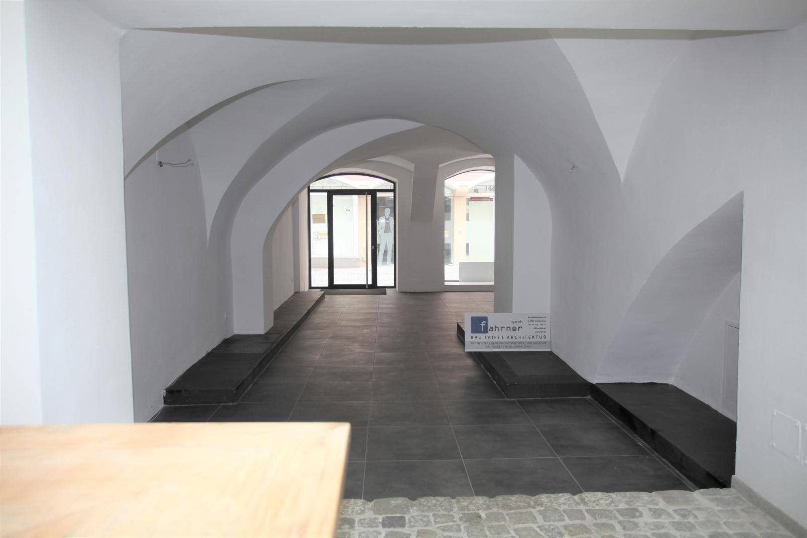 Langer Geschäftsraum mit Gewölbedecken, dunklem Fliesenboden und einer modernen Glastür zum Eingangsbereich.