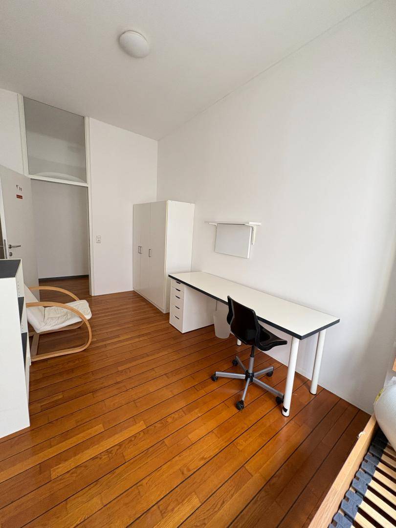Schlafzimmer mit Holzboden, Schreibtisch und Kleiderschrank, offen zum Flur.