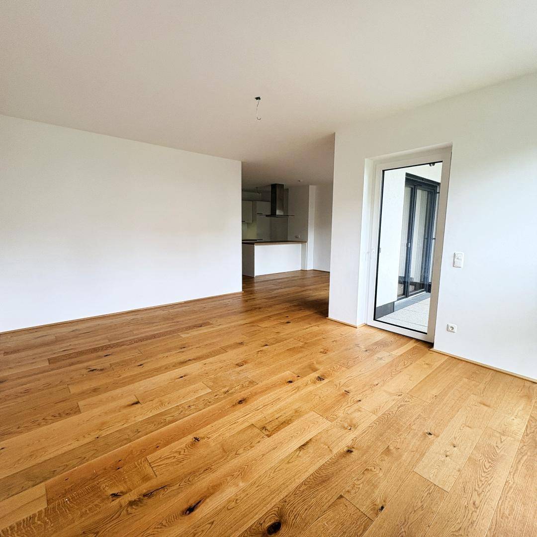 Geräumiger Wohnbereich mit hellem Holzboden und Zugang zu einem Außenbereich durch eine Glastür.