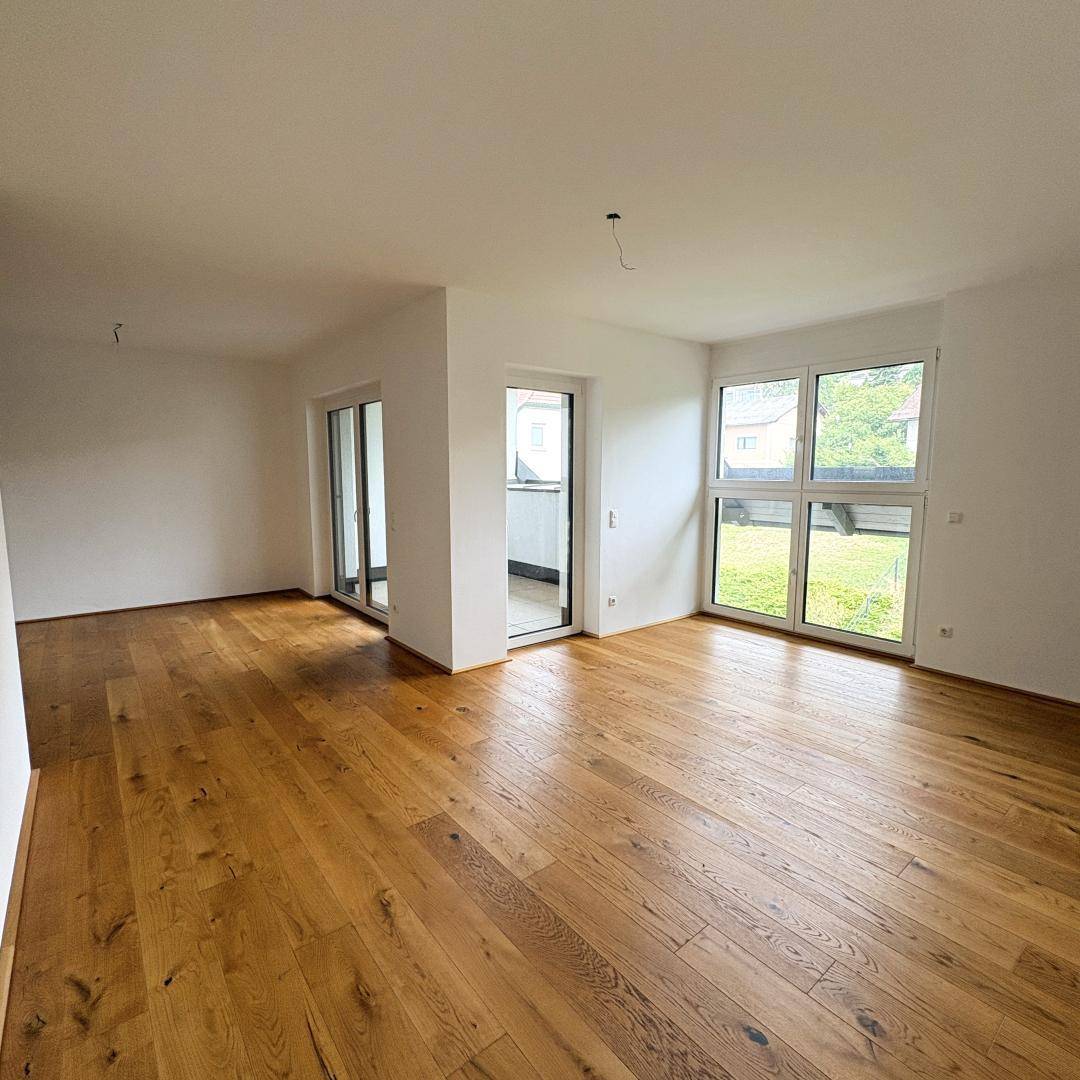 Heller Wohnbereich mit großen Fenstern, Holzboden und Zugang zu einem Balkon oder einer Terrasse.