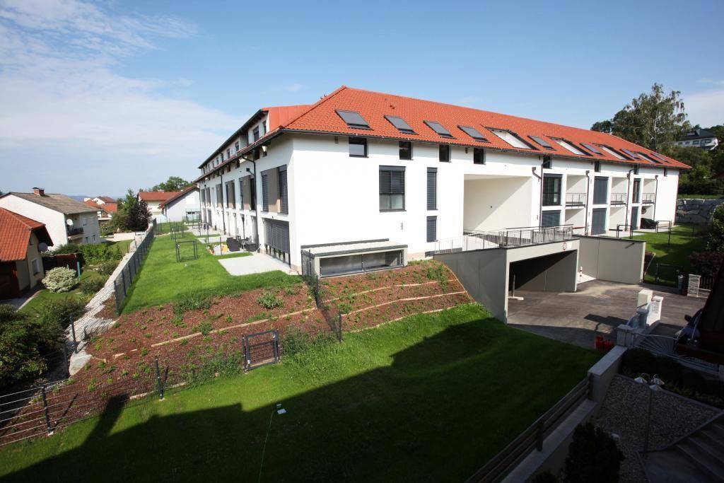 Neubau-Wohnanlage mit rotem Ziegeldach, Terrassen und Tiefgaragenzufahrt in grüner Umgebung.