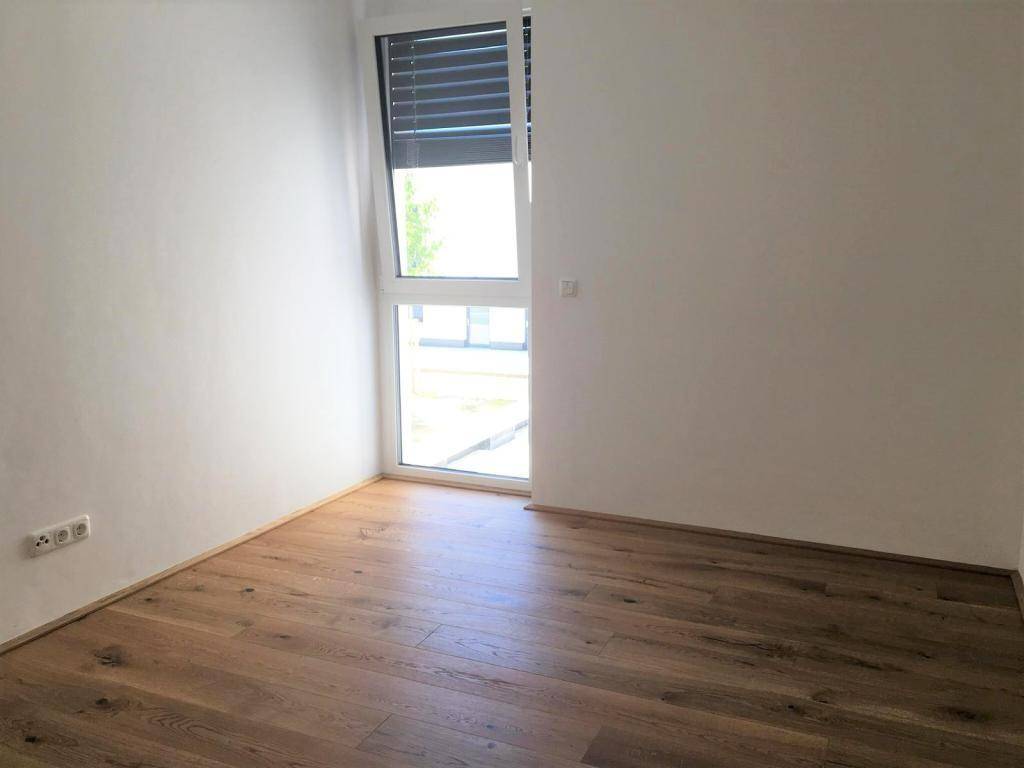 Helles Zimmer mit Holzboden und einem großen Fenster, das mit einem Rollladen ausgestattet ist.