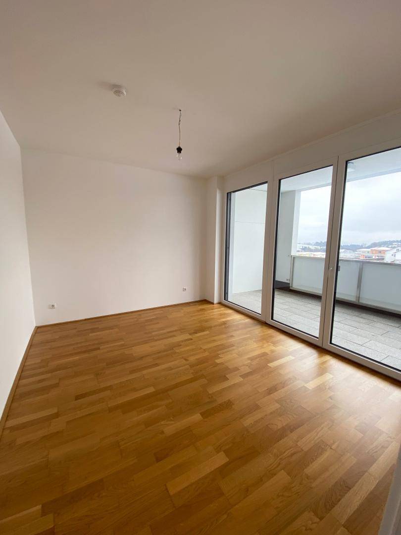 Heller Raum mit Parkettboden und großen Fenstern, die Zugang zu einem Balkon bieten.