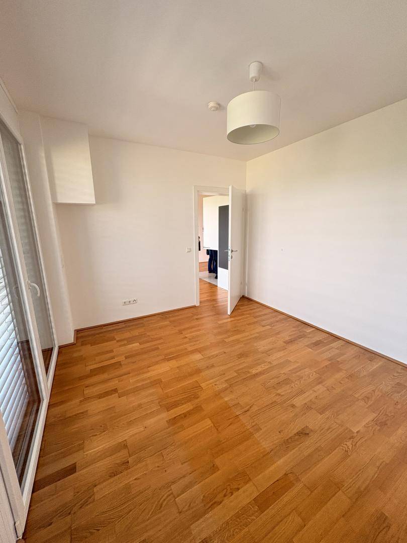 Leeres Schlafzimmer mit Parkettboden, weißen Wänden und einem Fenster mit Jalousien.