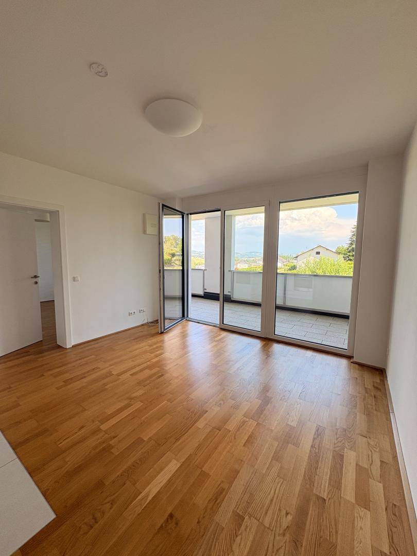 Geräumiges Wohnzimmer mit hellem Parkettboden und großen Fenstern, die auf einen Balkon führen.