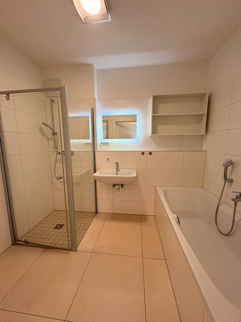 Modernes Badezimmer mit weißen Fliesen, Duschkabine, Badewanne und beleuchtetem Spiegel.