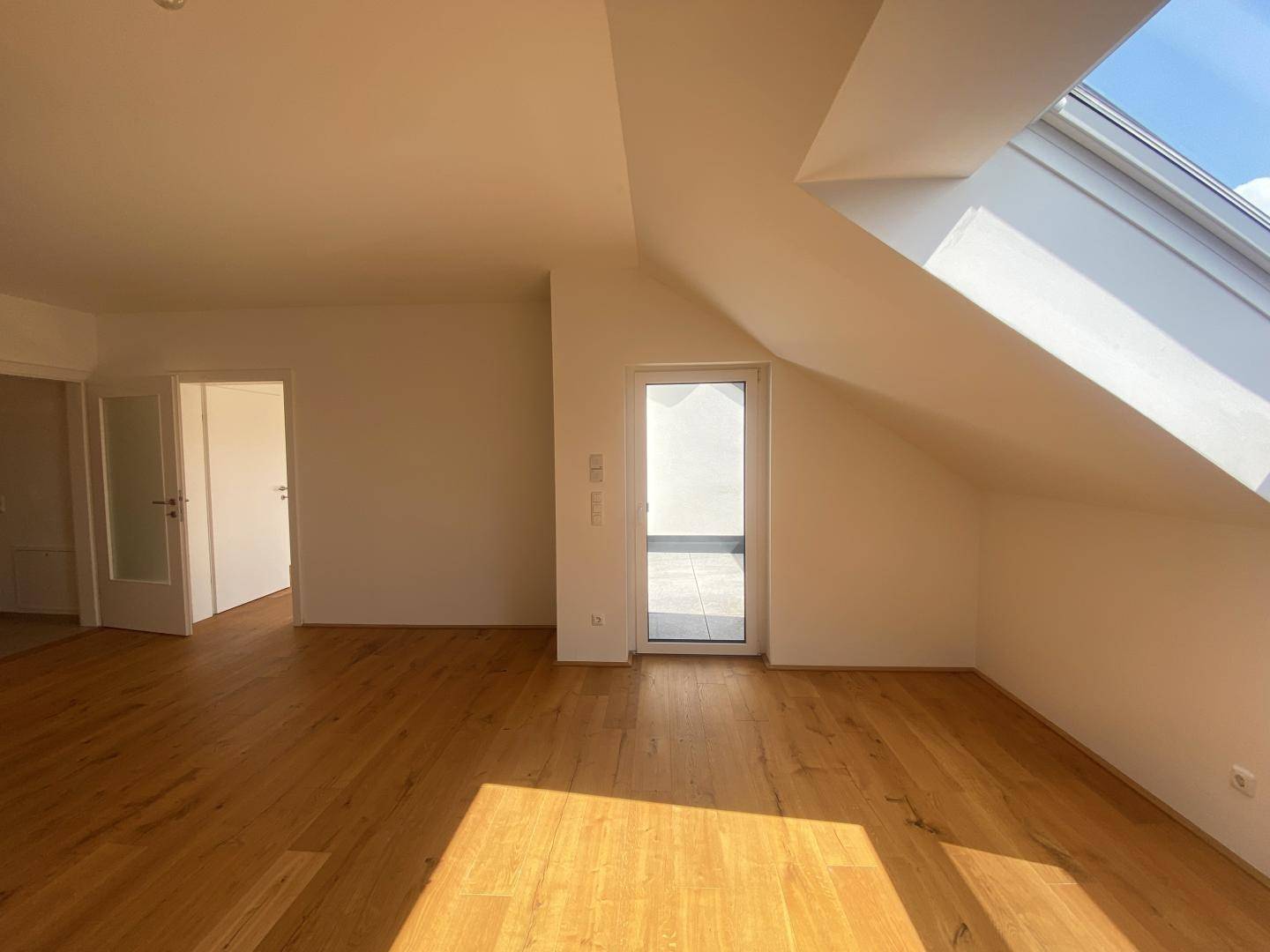 Lichtdurchfluteter Raum mit Holzboden, Dachschräge, großem Dachfenster und Zugang zur Loggia.