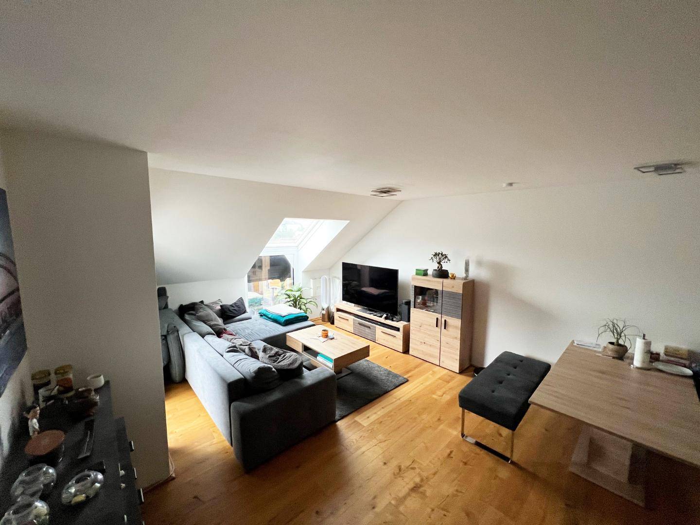 Gemütliches Wohnzimmer mit großem Sofa, Holzboden und Dachschräge, beleuchtet durch ein Dachfenster.