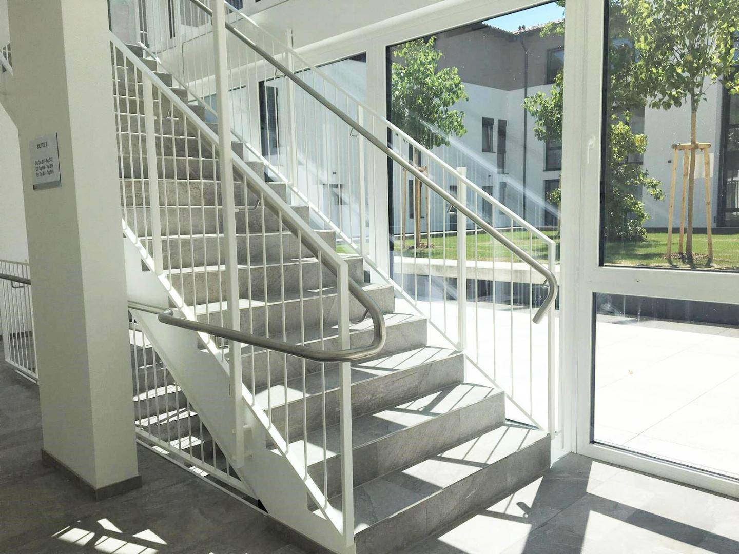Helles Treppenhaus mit modernen Geländern und großen Fenstern, die viel Tageslicht hereinlassen.