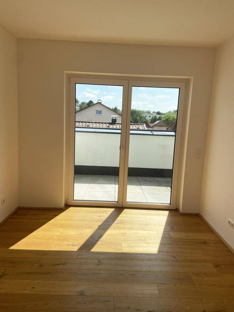 Helles Zimmer mit Holzboden und großer Glastür, die zu einem sonnigen Außenbereich führt.