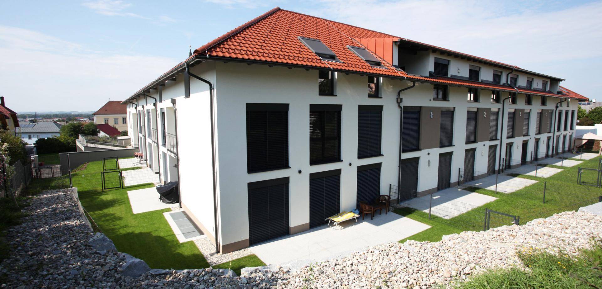 Modernes Gebäude mit rotem Ziegeldach, weißer Fassade und schwarzen Fensterläden, umgeben von Grünflächen.