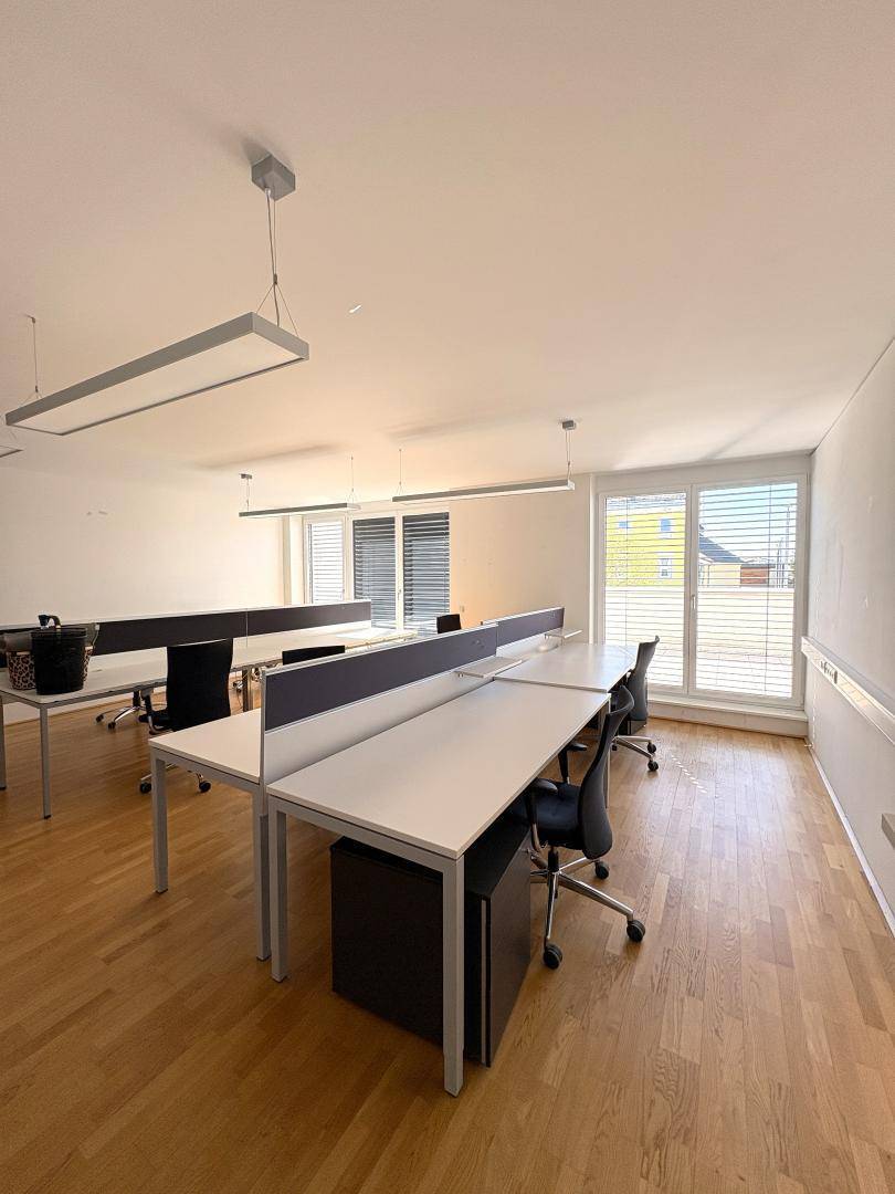 Moderner Büroraum mit mehreren Arbeitsplätzen, Holzboden und großen Fenstern mit Jalousien.
