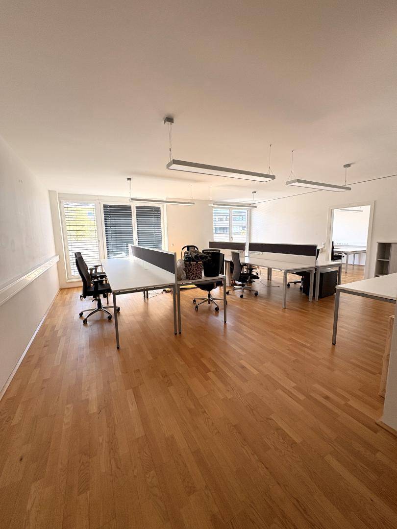 Großes, offenes Büro mit mehreren Arbeitsplätzen, Holzboden und großen Fenstern mit Jalousien.