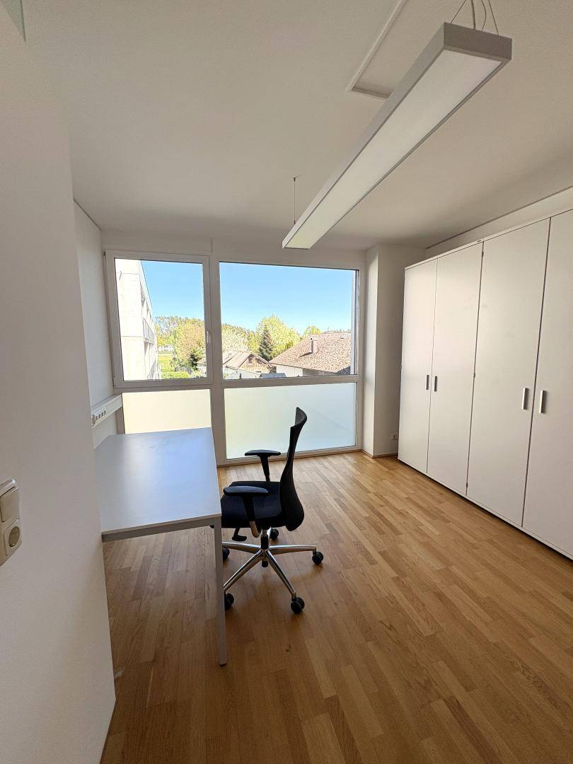 Geräumiges Büro mit Holzboden, großen Fenstern und Blick ins Grüne, ausgestattet mit einem Schreibtisch.