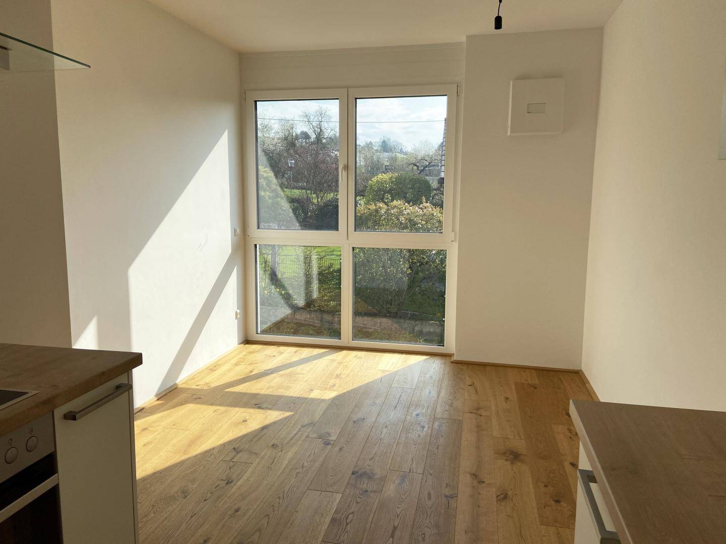 Heller Raum mit Holzboden und großem Fenster, das viel Tageslicht und einen Blick ins Grüne bietet.