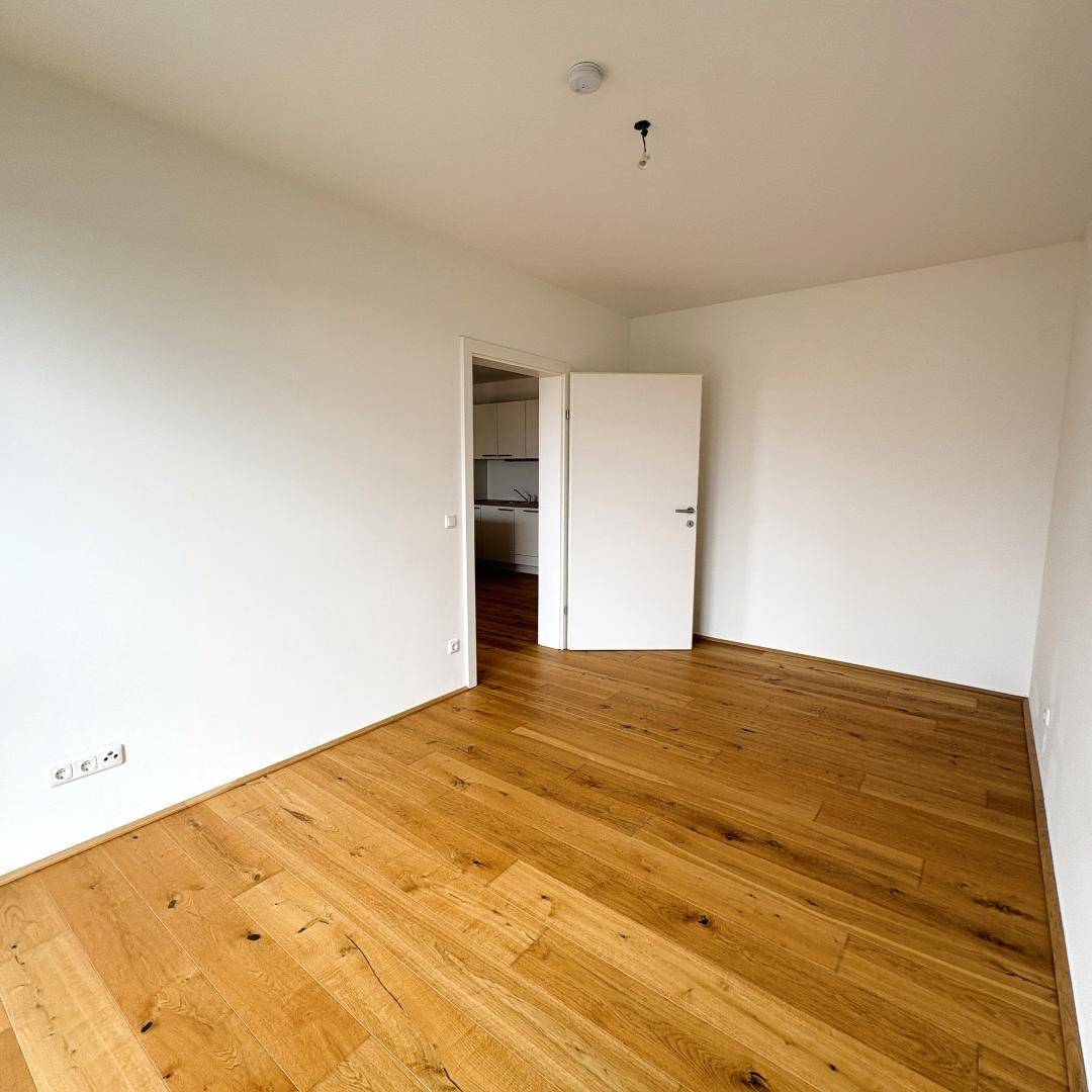 Leeres Zimmer mit Parkettboden und Zugang zu einem weiteren Raum, möglicherweise der Küche.
