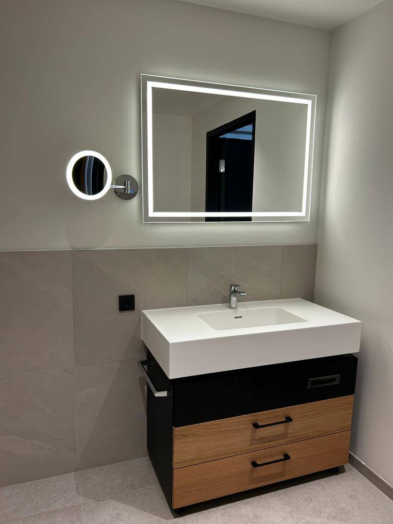 Modernes Badezimmer mit beleuchtetem Spiegel, Einzelwaschbecken und Holz-Waschtischunterschrank.