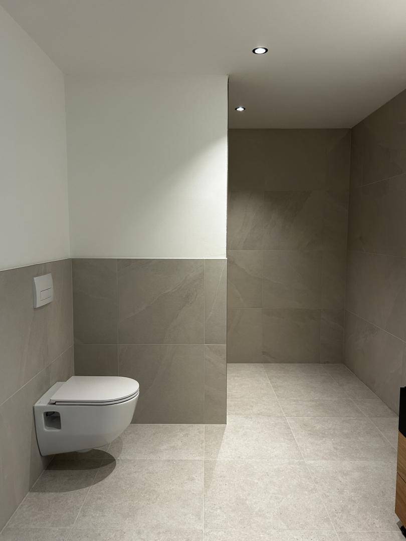 Geräumiges Badezimmer mit großen Fliesen, wandhängendem WC und begehbarer Dusche für hohen Komfort.
