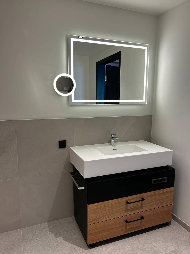Modernes Badezimmer mit beleuchtetem Spiegel, Einzelwaschbecken und Holzunterschrank auf Fliesenboden.