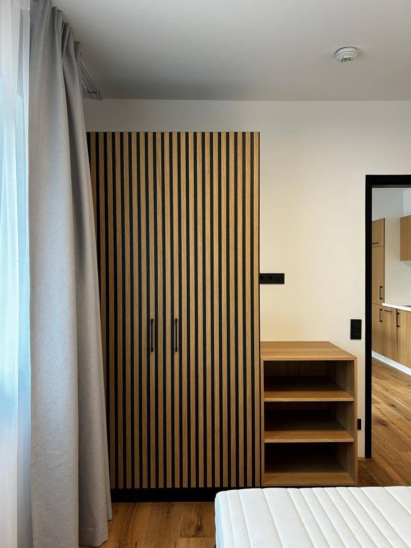 Moderner Kleiderschrank mit Holzlamellen und integriertem Regal im Schlafzimmerbereich, neben einem Fenster.