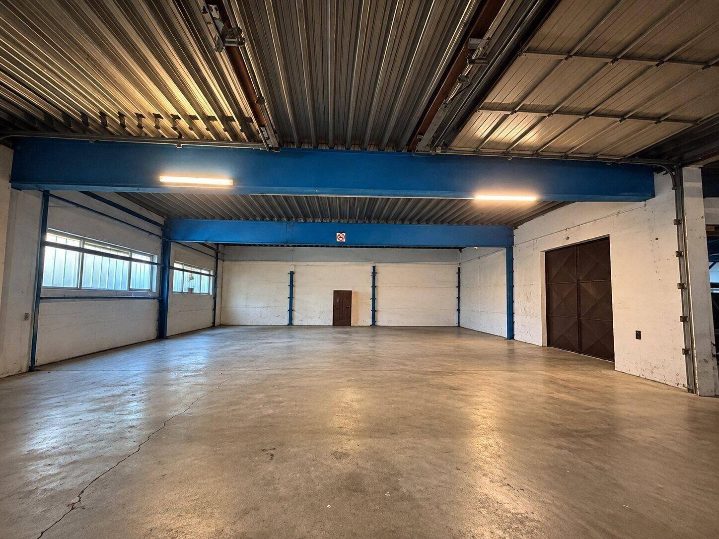 Leere Lagerhalle mit Betonboden, blauen Stahlträgern und mehreren Fenstern für gute Belüftung und Licht.