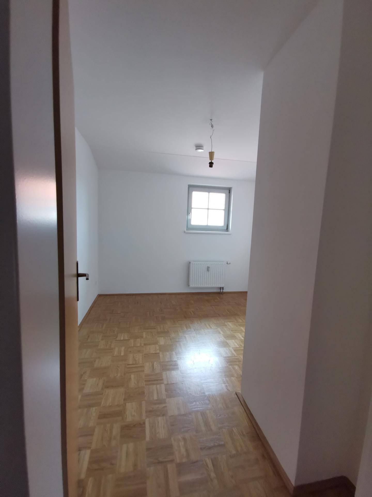 Blick in einen leeren Raum mit Parkettboden, weißen Wänden und einem Fenster mit Heizkörper.