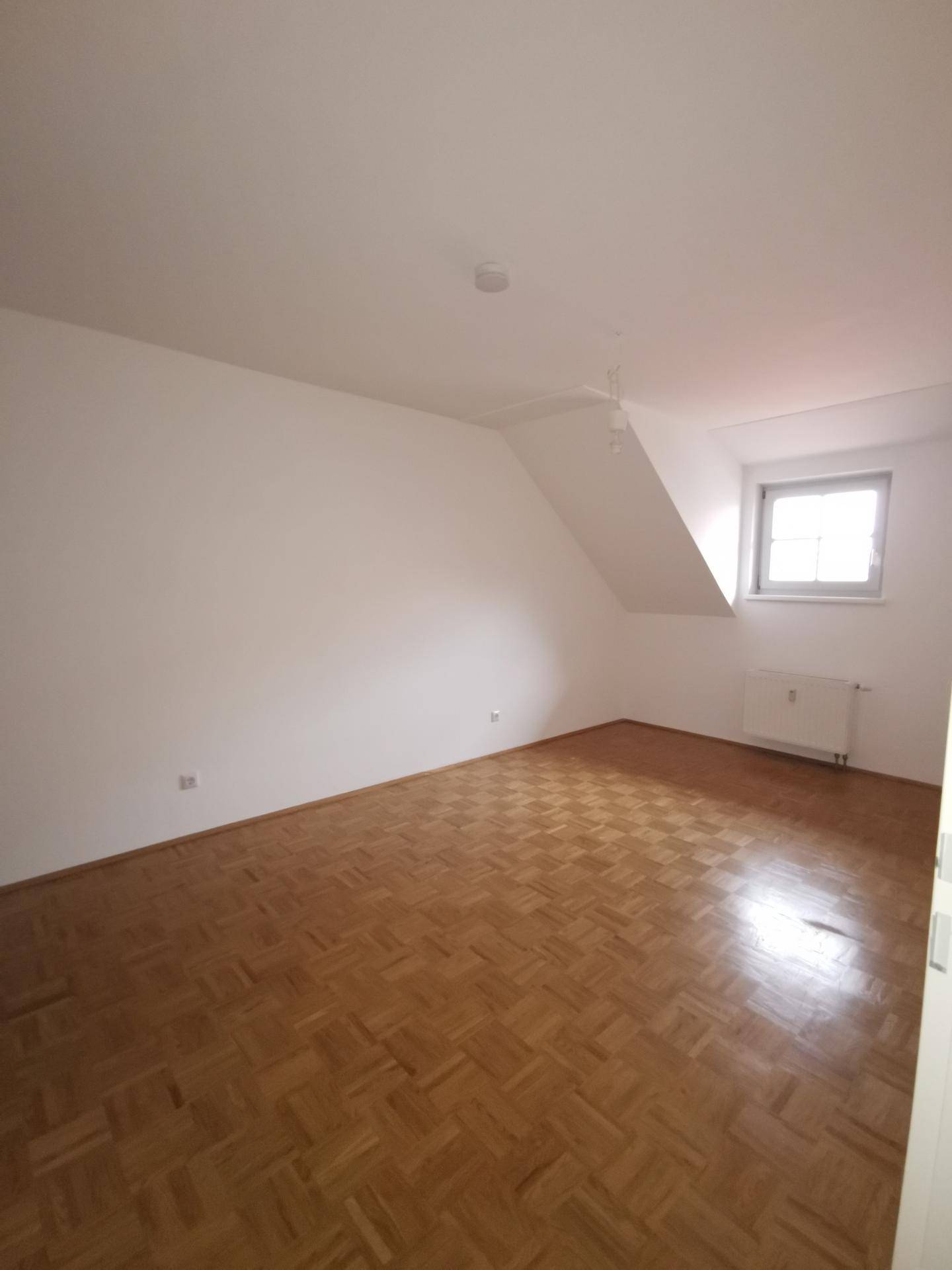 Geräumiges Schlafzimmer mit Parkettboden, Dachschräge und einem Fenster, das für natürliche Belichtung sorgt.