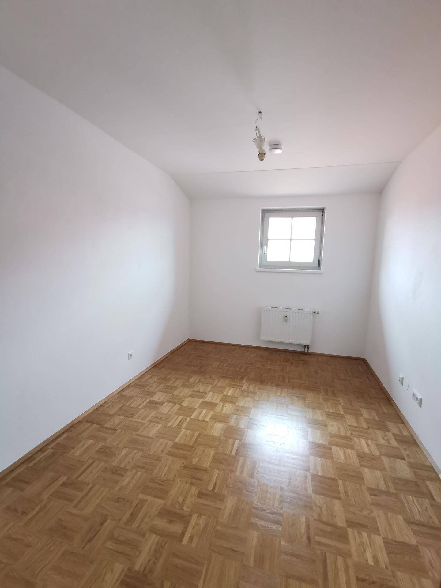 Helles Schlafzimmer mit Parkettboden, weißen Wänden und einem Fenster, das Tageslicht hereinlässt.