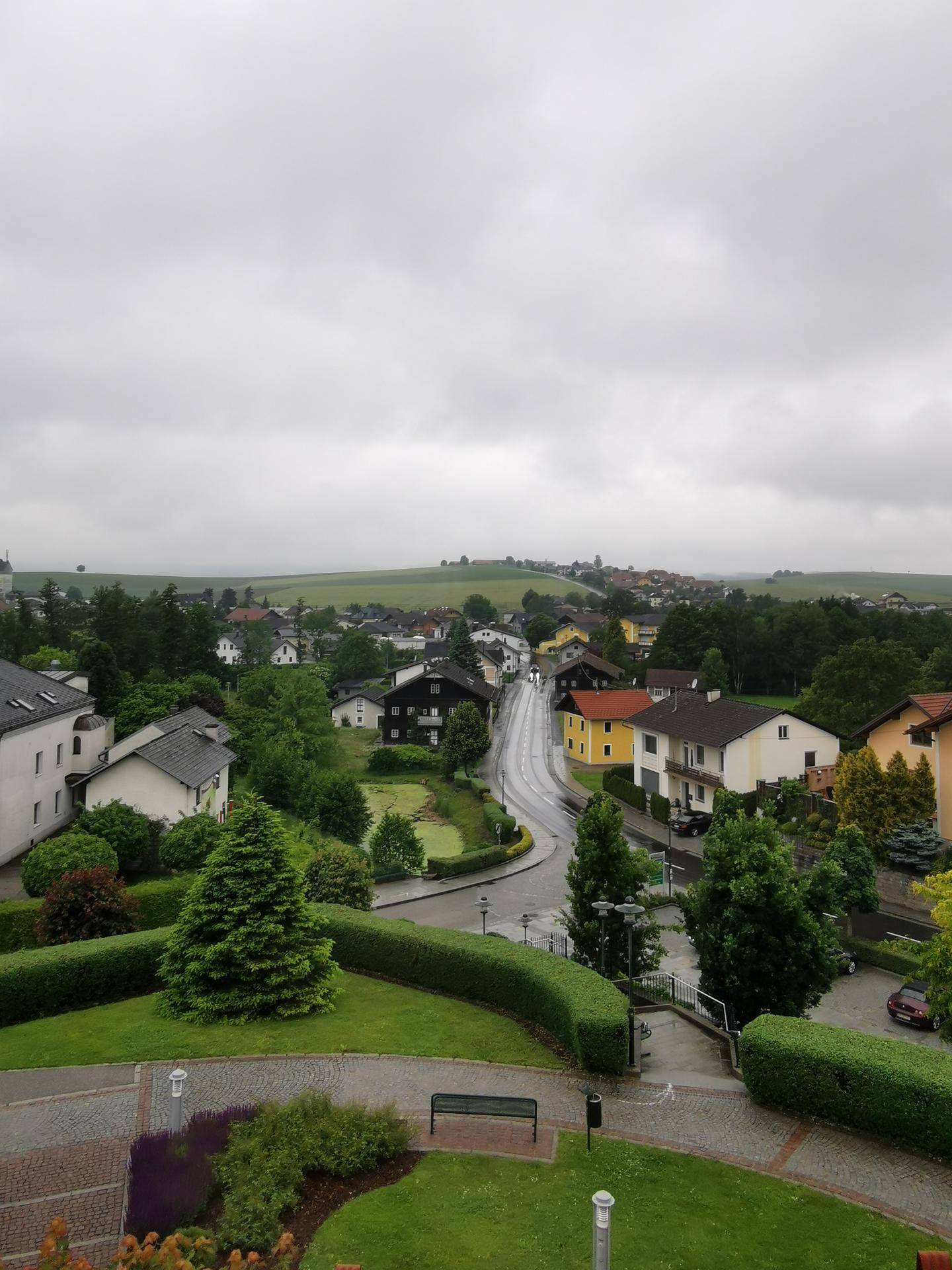Panoramablick über eine ländliche Siedlung mit Häusern, viel Grün und einer Straße unter bewölktem Himmel.