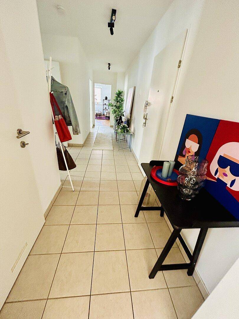 Heller Flur mit Fliesenboden, Garderobe und einem kleinen Tisch mit farbenfrohem Kunstwerk.